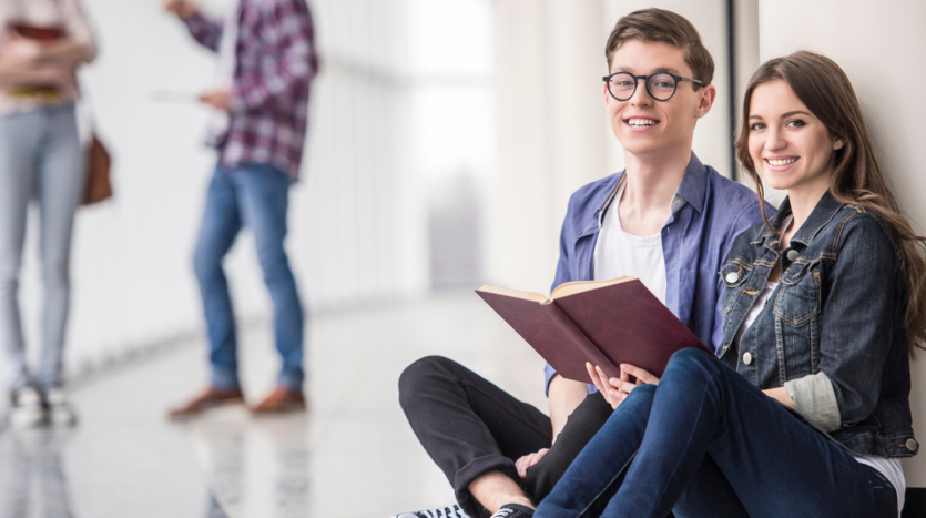 Un chico y una chica jóvenes sentados en el suelo del pasillo del colegio mirando a cámara y sonriendo mientras sujetan un cuaderno