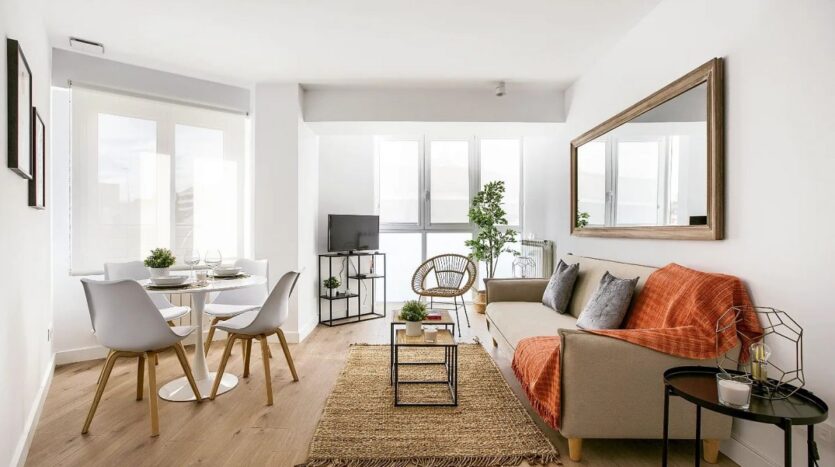 Fotografía de salón con una mesa con dos sillas blancas a la izquierda, una mesita marrón en medio encima de la alfombra marrón y un sofá marrón con manta naranja a la derecha y arriba de este un espejo. Los ventanales dejan entrar luz natural y hay una televisión al fondo y a su derecha una planta verde.